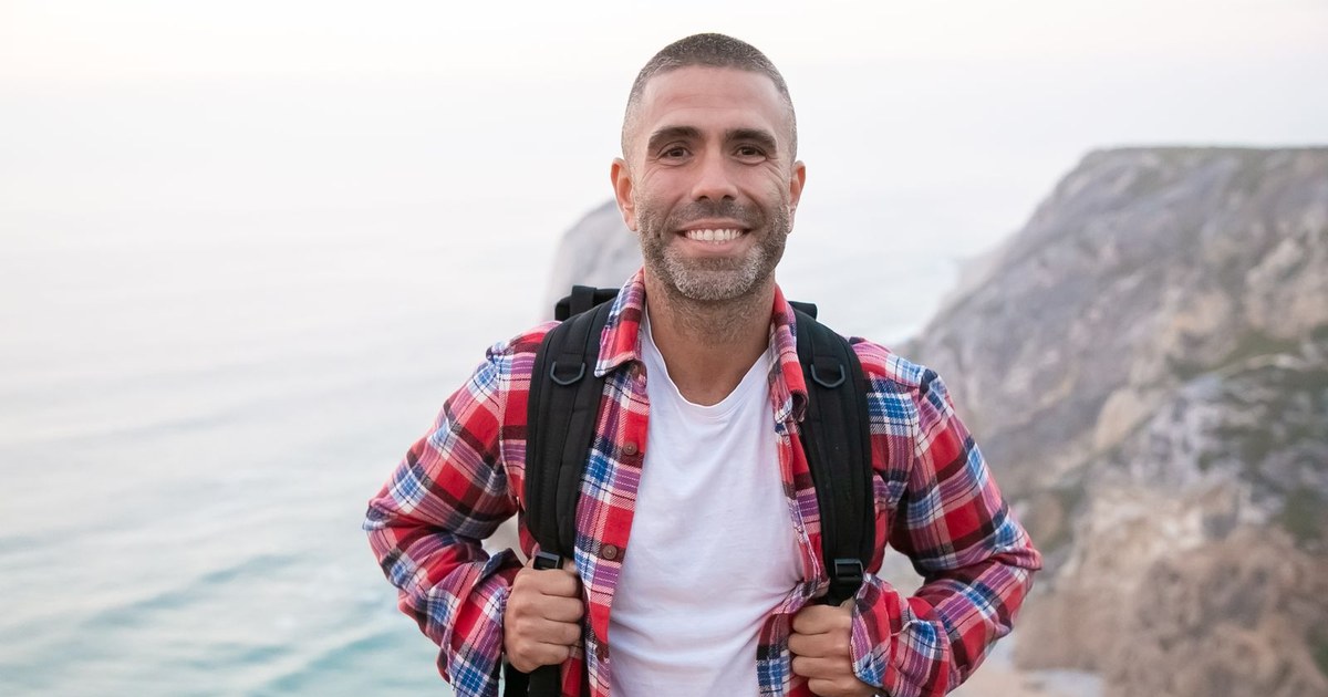 A healthy, smiling adult man in a plaid shirt hiking outdoors on sunlit coastal cliffs, conveying vitality and well-being