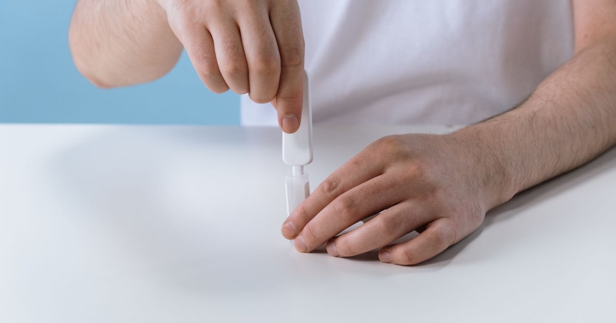 A person's hands using a dropper and collection tube to perform an at-home self-test on a white table