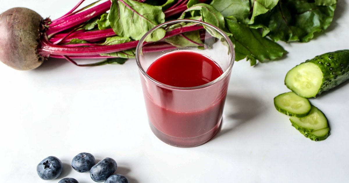 A glass of deep-red beet juice beside a whole beetroot with leafy green tops, cucumber slices and blueberries on a white surface