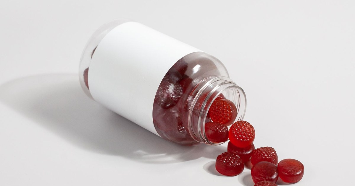 Deep red gummies spilling from a tipped clear bottle with a blank white label on a white surface