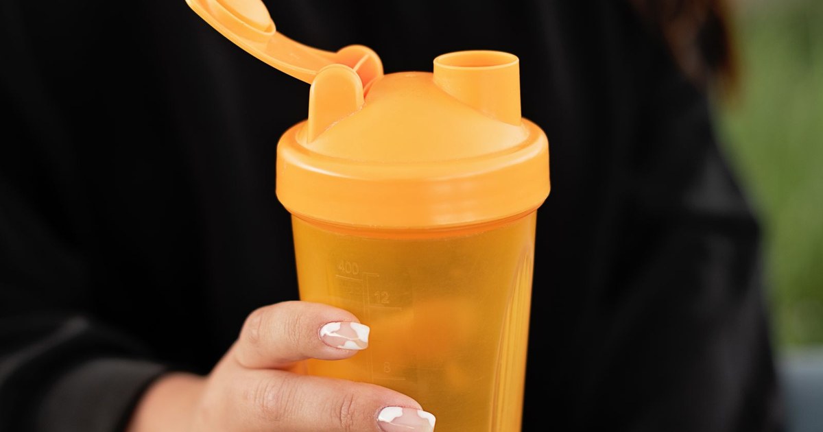 A hand holding a bright orange protein shaker bottle outdoors