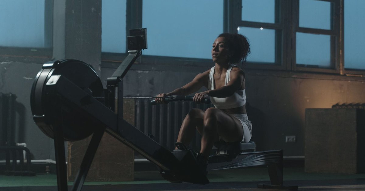 A woman in white workout clothes pulling hard on an indoor rowing machine in a dim gym with tall windows