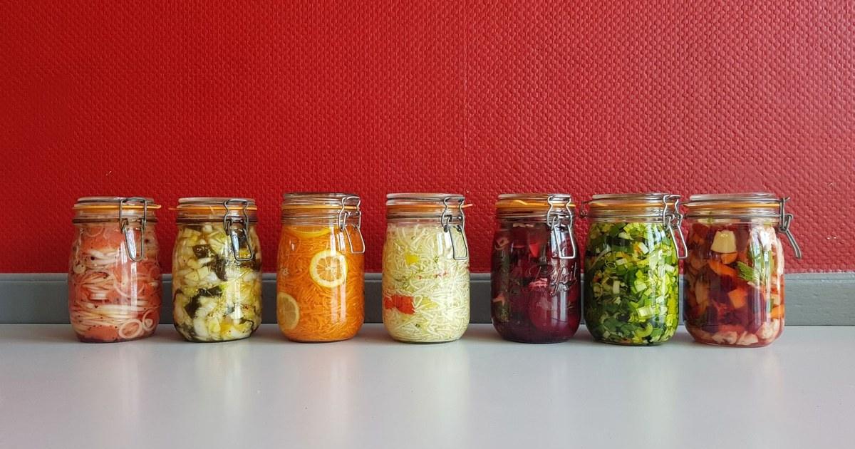 A row of glass jars of colorful fermented vegetables against a red wall, the fermented foods that feed the gut bacteria that make butyrate