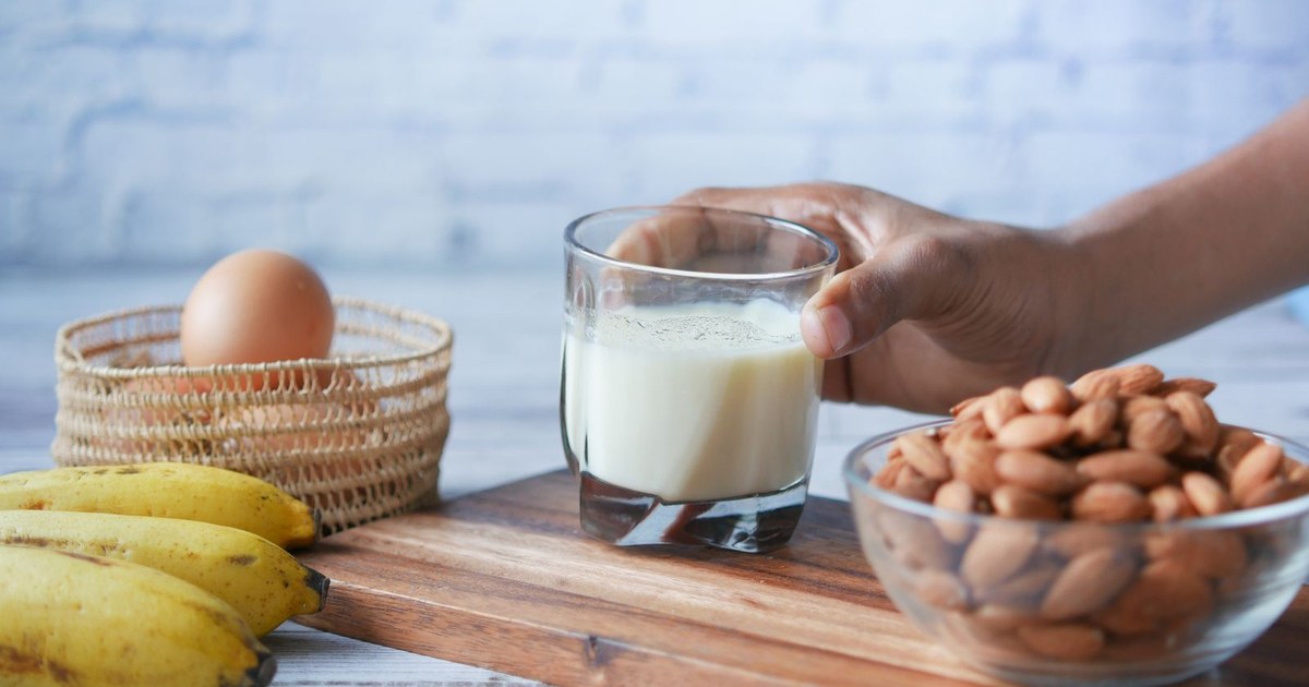 A hand reaching for a glass of milk beside a bowl of almonds, an egg, and bananas on a wooden board