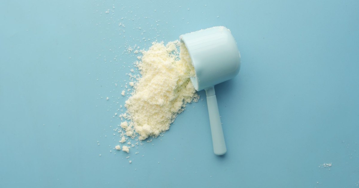 A pale blue plastic supplement scoop tipped on its side with white creatine powder spilling into a small pile on a light blue background