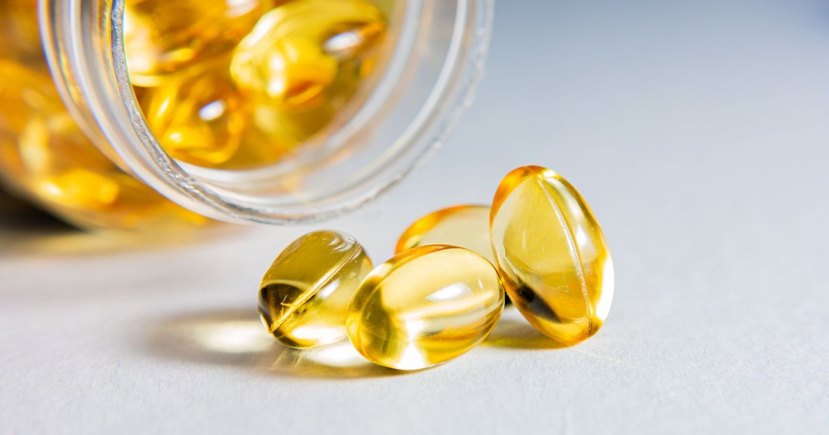 Glossy golden-amber softgel capsules spilling from a clear glass jar onto a bright white and pale blue surface