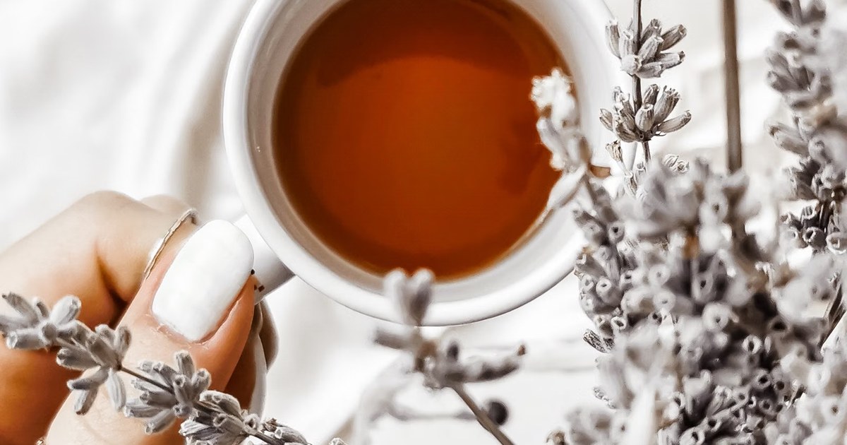 A hand holding a warm cup of tea among sprigs of dried lavender on a soft white surface