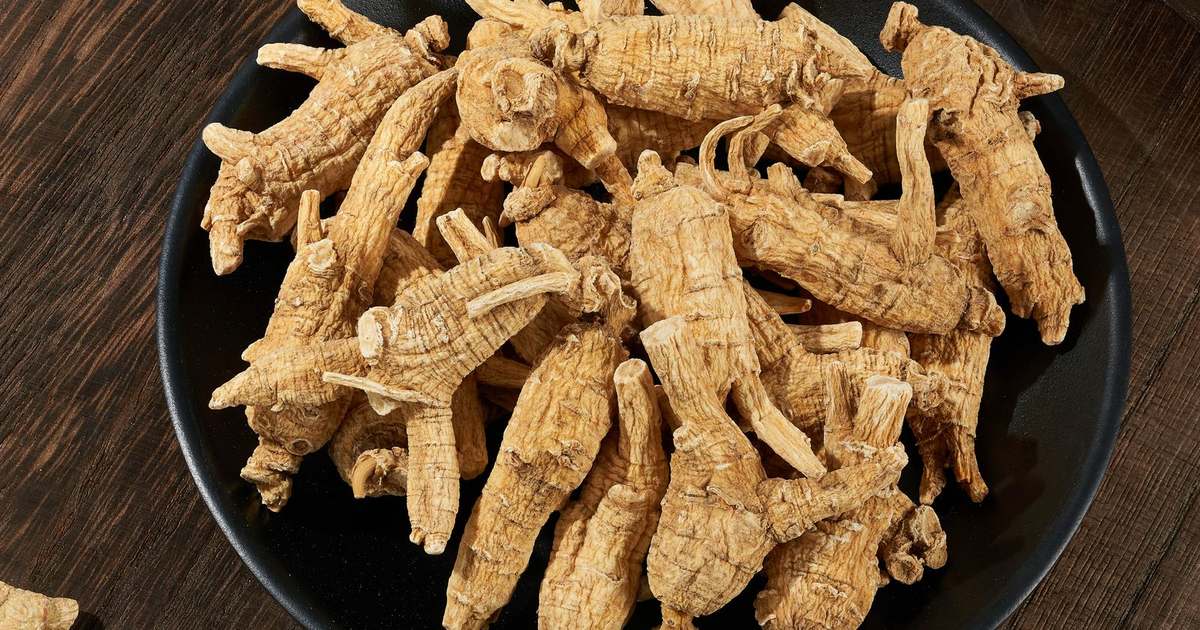 Dried ginseng roots piled in a black ceramic bowl on a dark wooden table with a few sliced pieces on a bamboo spoon nearby
