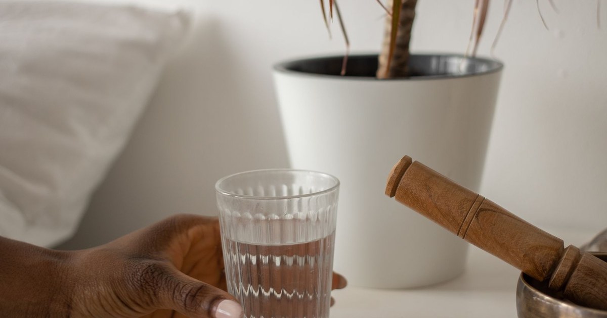 A hand setting a ridged glass of water on a white bedside table beside a potted plant and a singing bowl