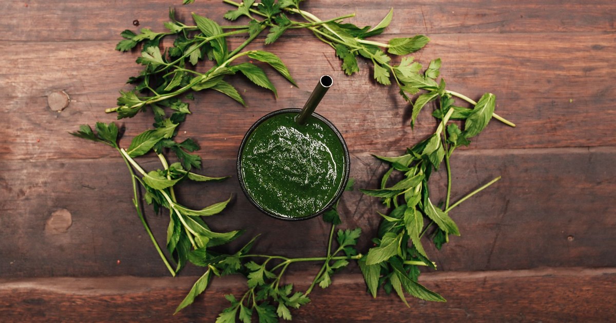 A green smoothie in a cup surrounded by fresh greens on a wooden table