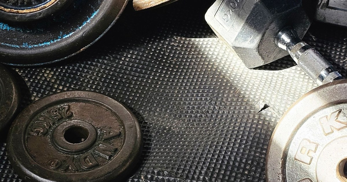 Worn iron weight plates and a chrome dumbbell on a textured rubber gym floor in a shaft of light