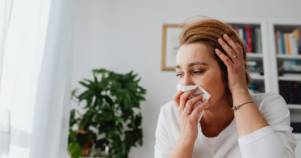 A woman blowing her nose into a tissue at home while feeling unwell with a cold