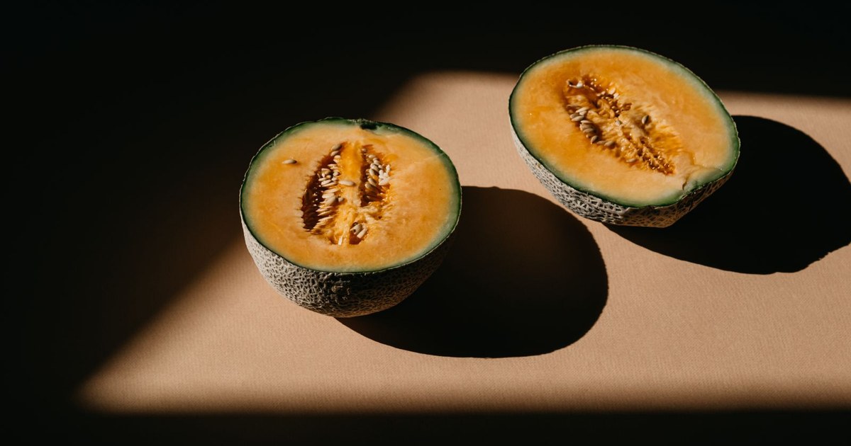 Two cantaloupe halves casting long shadows on a beige surface in a band of sunlight