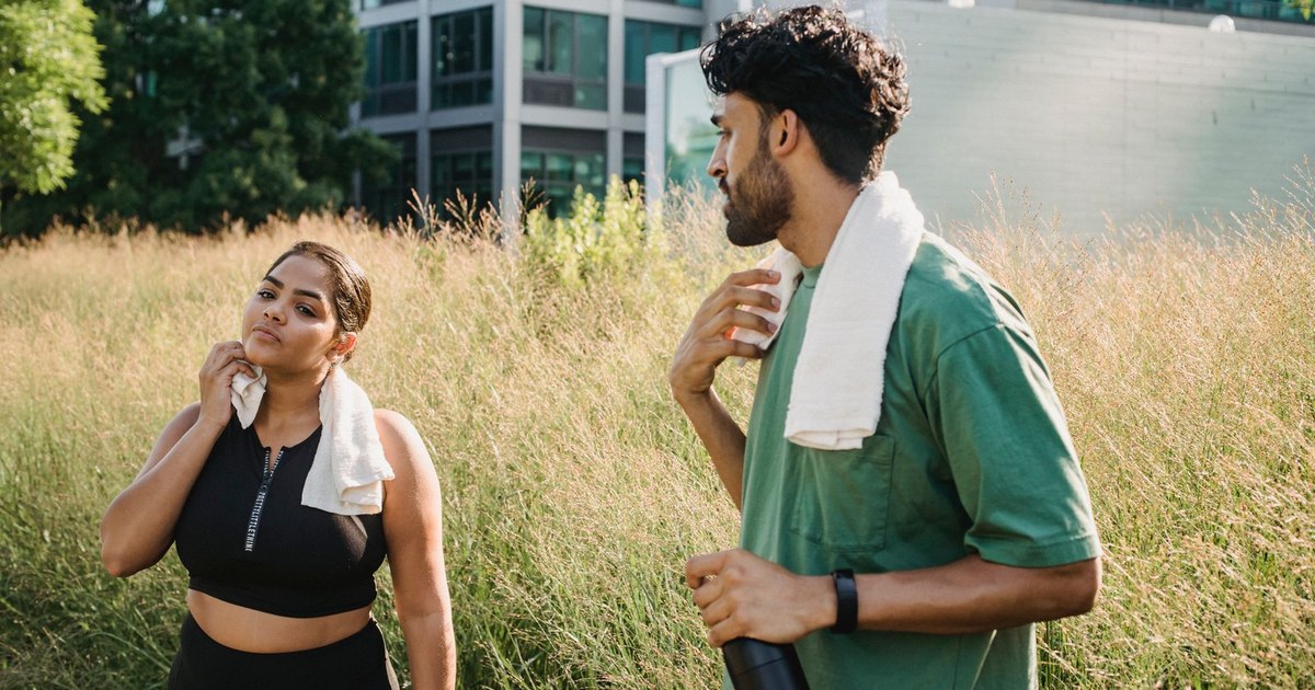 A woman and a man with towels around their necks cooling down after a workout in tall grass beside city buildings