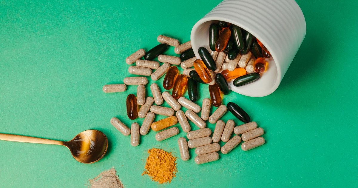 Assorted supplement capsules and amber softgels spilling from a white cup onto a green surface with a wooden spoon and a small mound of turmeric powder