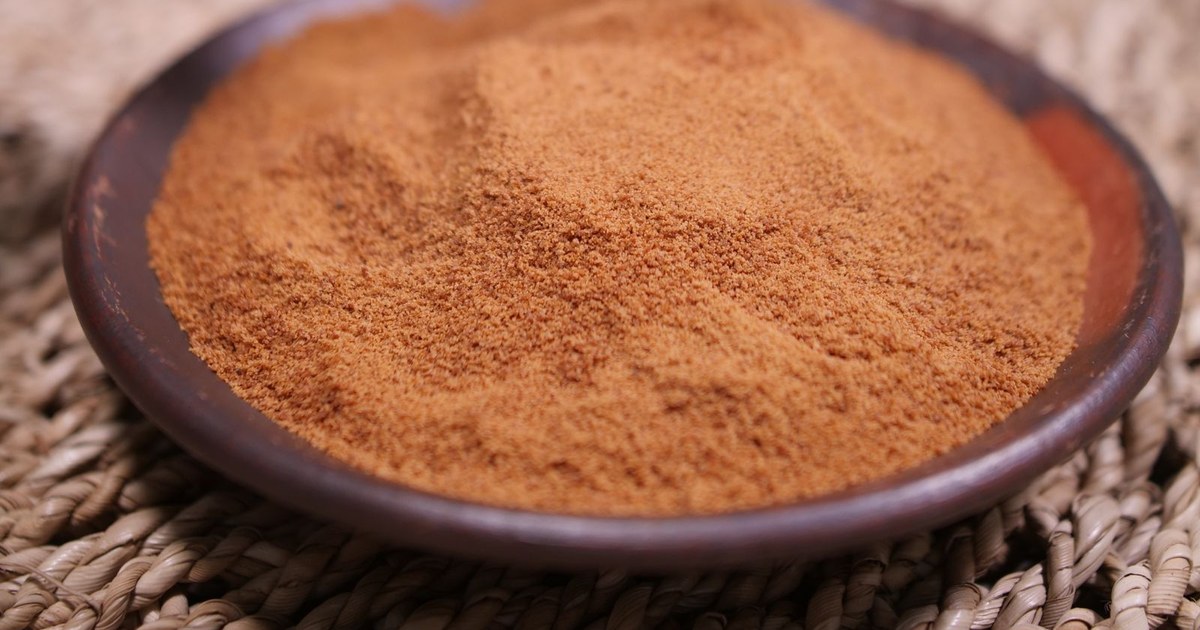 A round wooden dish filled with tan brown maca root powder resting on a woven straw mat