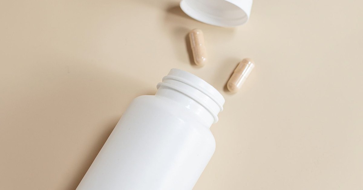 A white supplement bottle with its cap off and two tan capsules on a beige background