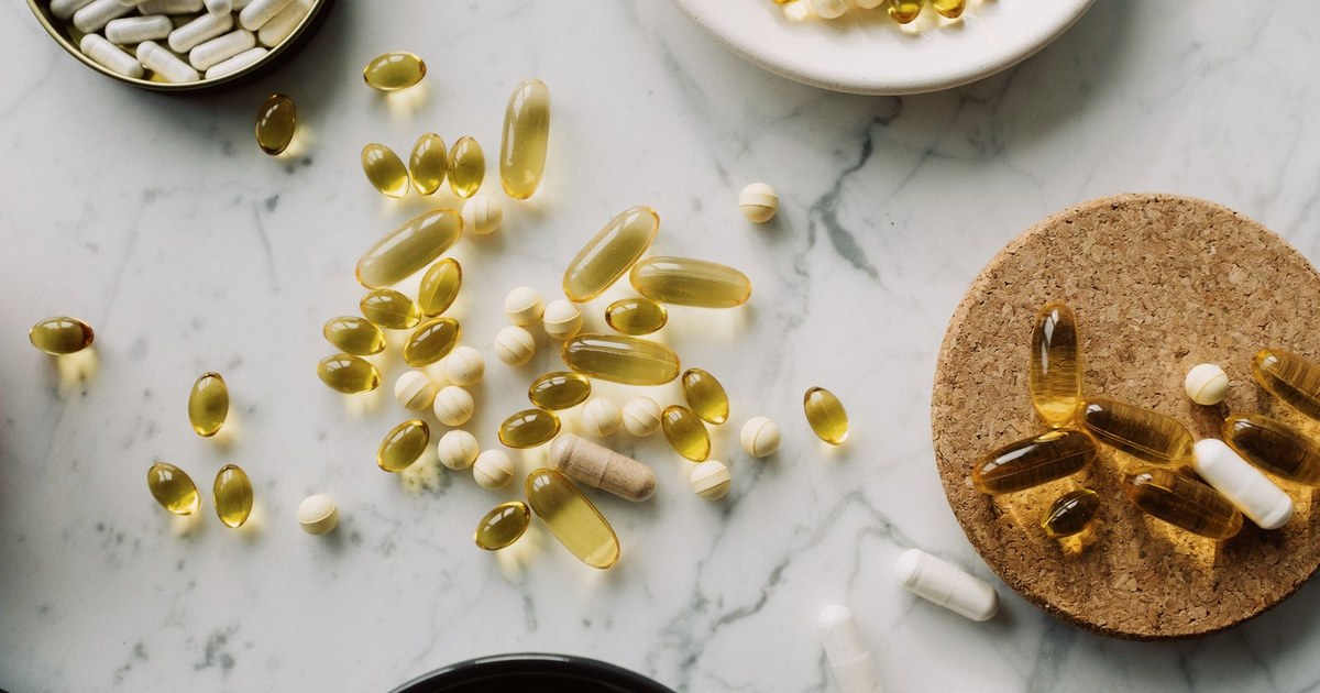 An overhead flat-lay of golden softgels and white capsules and tablets on a marble surface with a cork trivet and small dishes