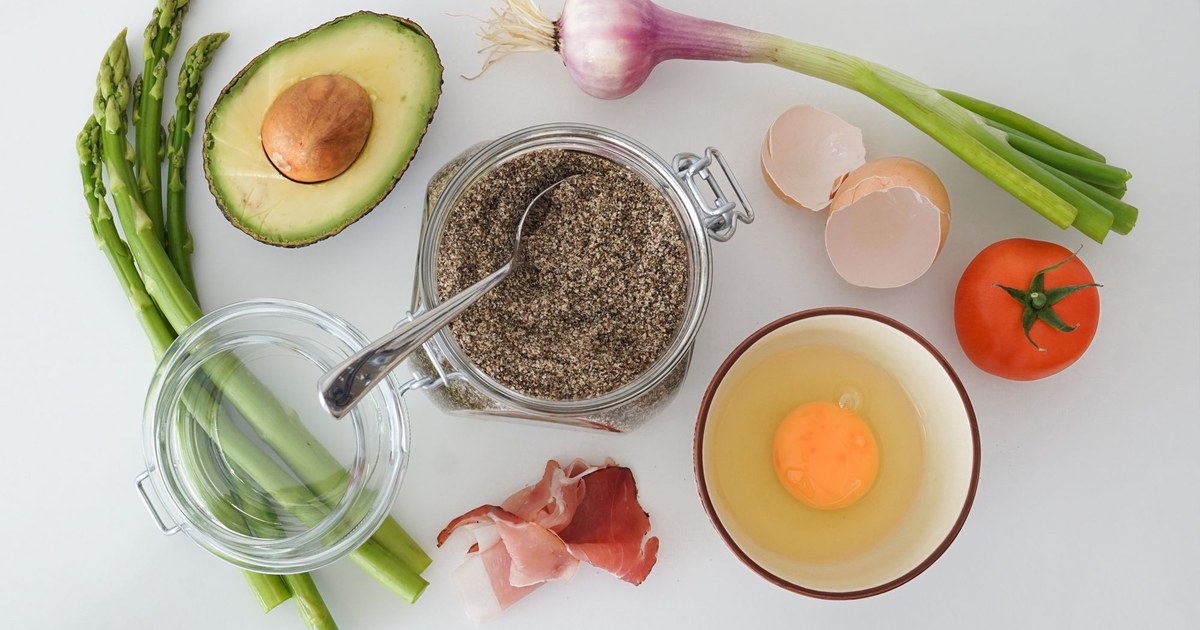 A flat lay of fresh whole foods including avocado, asparagus, egg, and tomato on a white surface