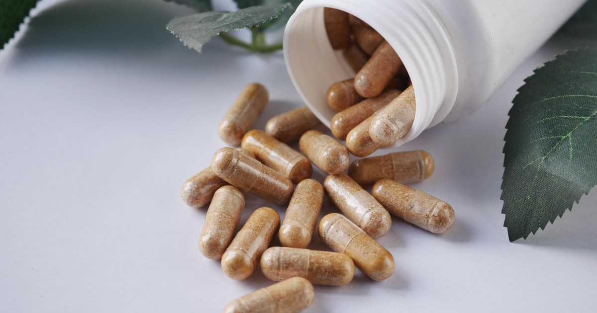 Beige supplement capsules spilling from a white bottle next to green leaves on a white surface