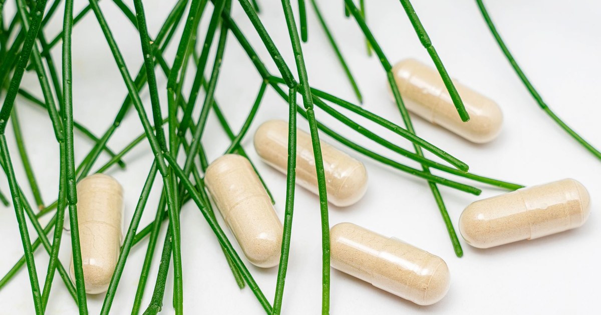 Beige supplement capsules scattered among fresh green plant stems on a white surface