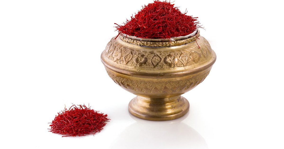 A brass bowl heaped with deep red saffron threads on a white background, with a small pile of threads beside it