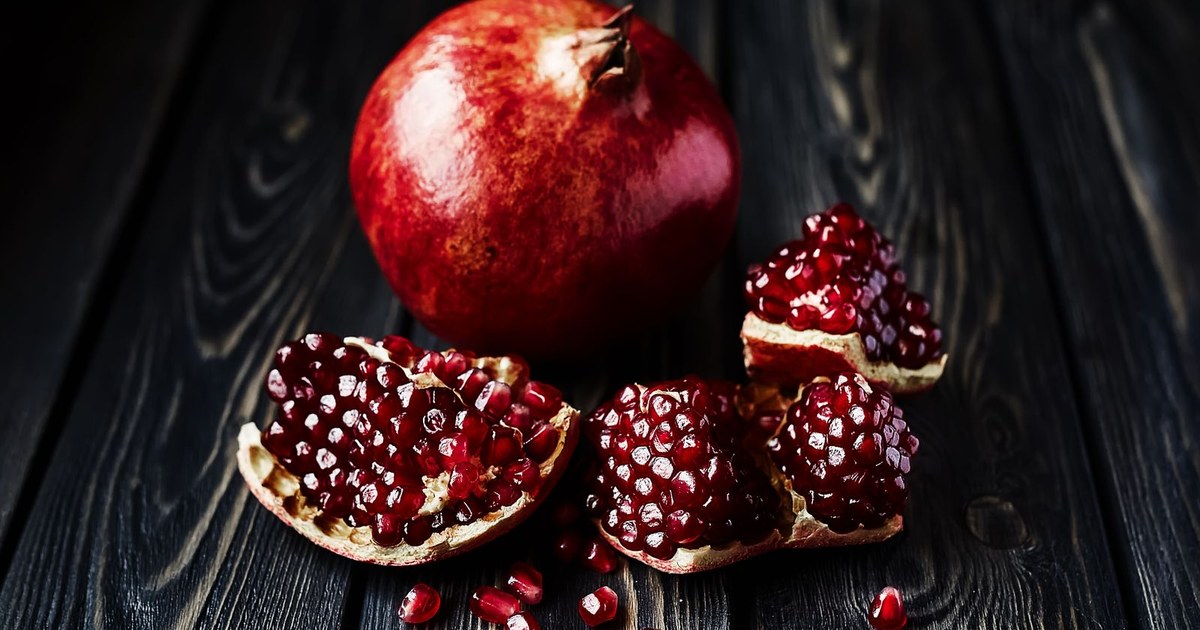 A whole pomegranate and broken pieces with scattered red seeds on a dark wooden table
