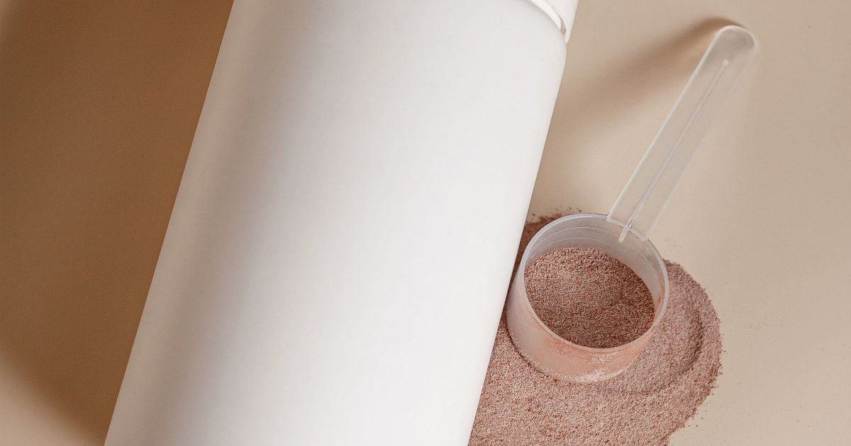 A white protein powder tub beside a scoop of tan plant protein powder spilling onto a warm beige surface