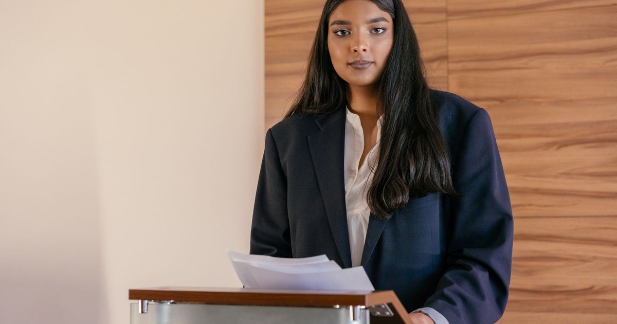 A composed woman in a dark navy blazer stands at a wooden lectern holding a few sheets of notes, looking calmly toward the audience before speaking, with a warm wood-panel wall behind her