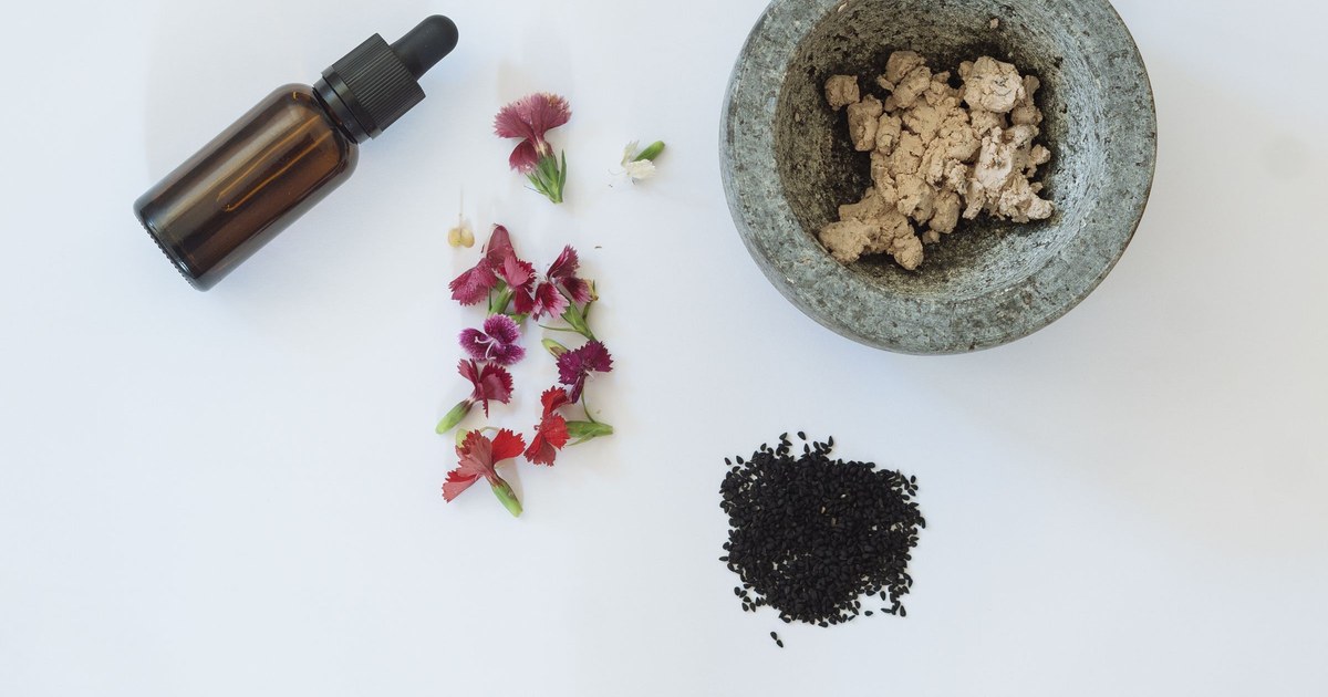 A flat-lay of an amber dropper bottle of black seed oil with Nigella sativa (black cumin) seeds, flowers, and a stone mortar