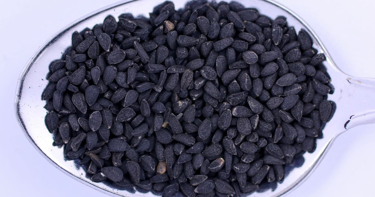 A close-up overhead shot of a metal spoon heaped with small matte-black teardrop-shaped Nigella sativa black cumin seeds on a pale background