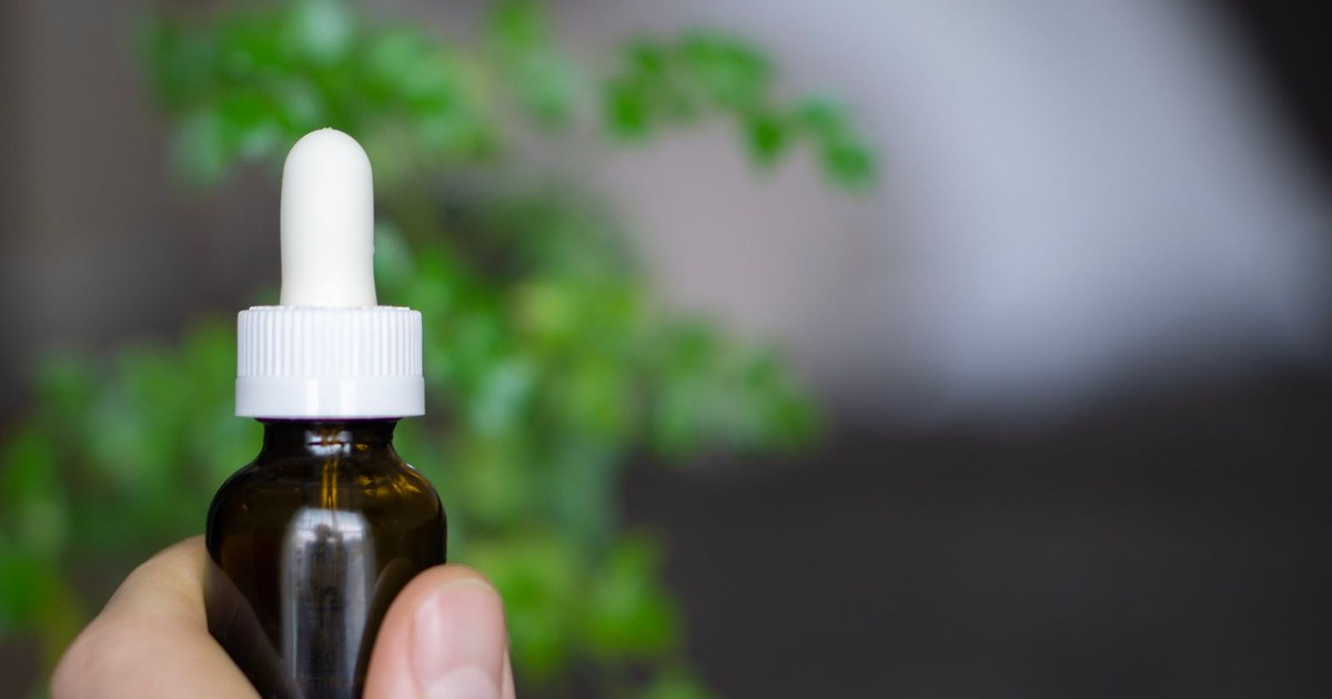 A hand holding an unlabeled amber glass dropper bottle of CBD oil in front of soft green foliage in natural light
