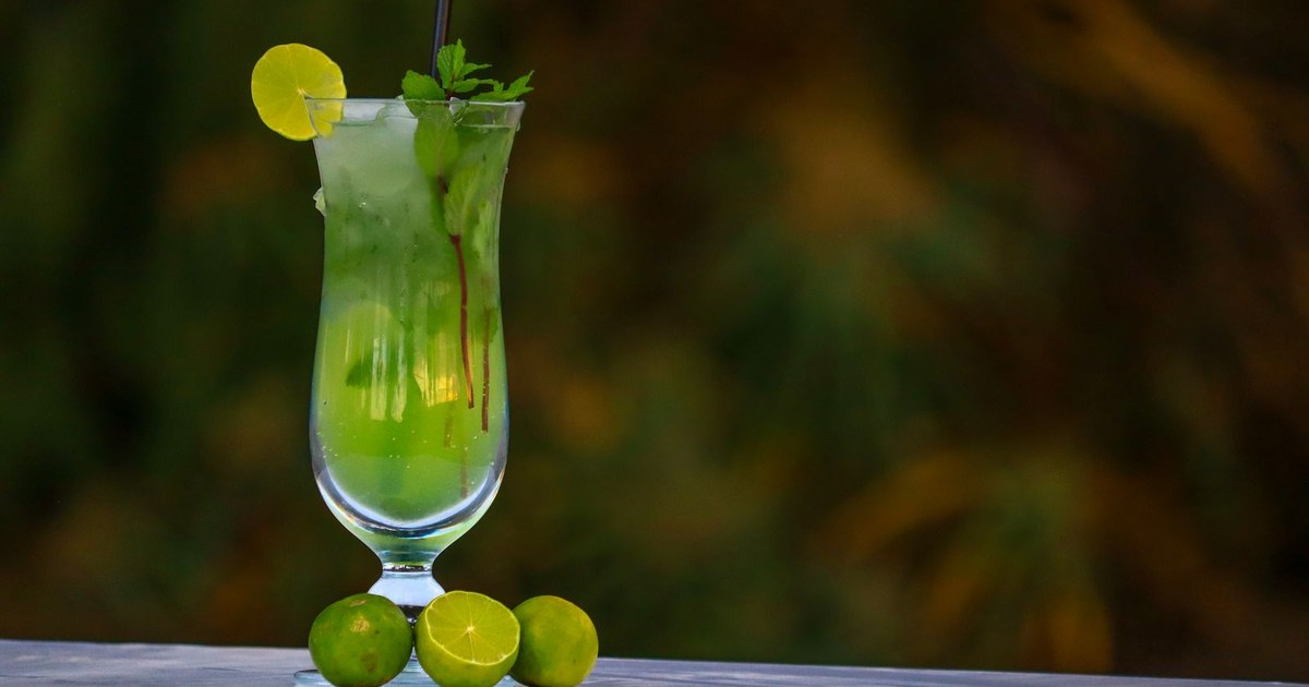 Tall glass of translucent pale green lime and mint drink with ice, a lime wheel, and whole limes on a table