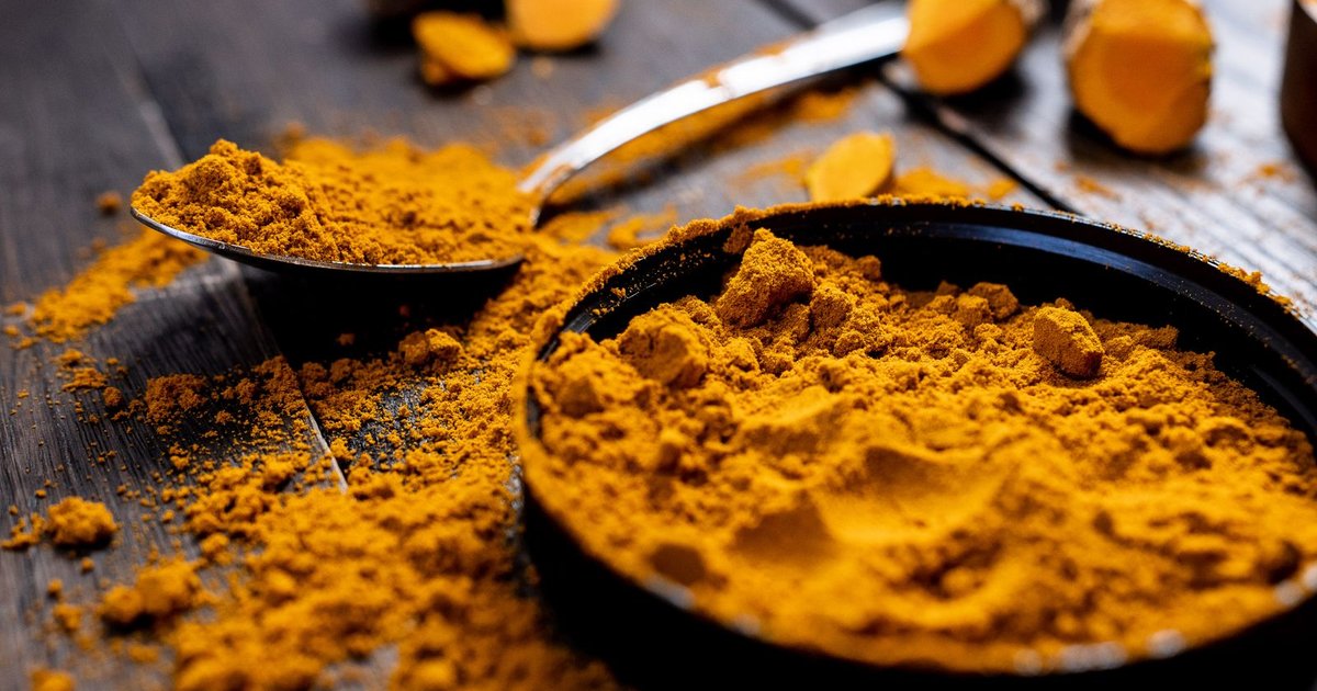 A dark round bowl heaped with vibrant golden-orange turmeric powder beside a metal spoon of the same powder, with loose powder scattered across a rustic dark-wood table and cut pieces of fresh turmeric root in the background