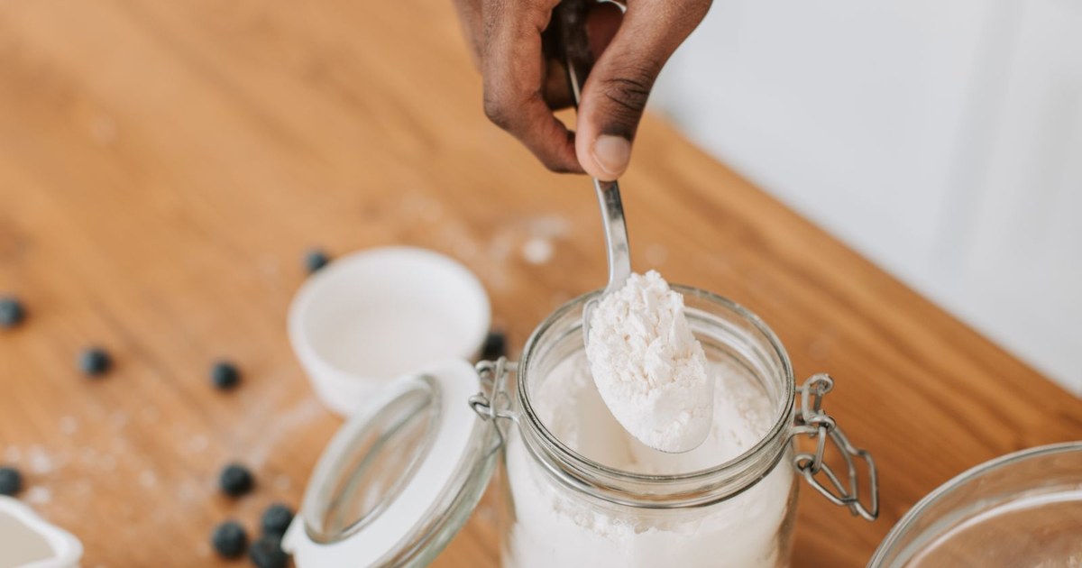 A hand lifting a spoonful of white powder from an open glass jar on a wooden table