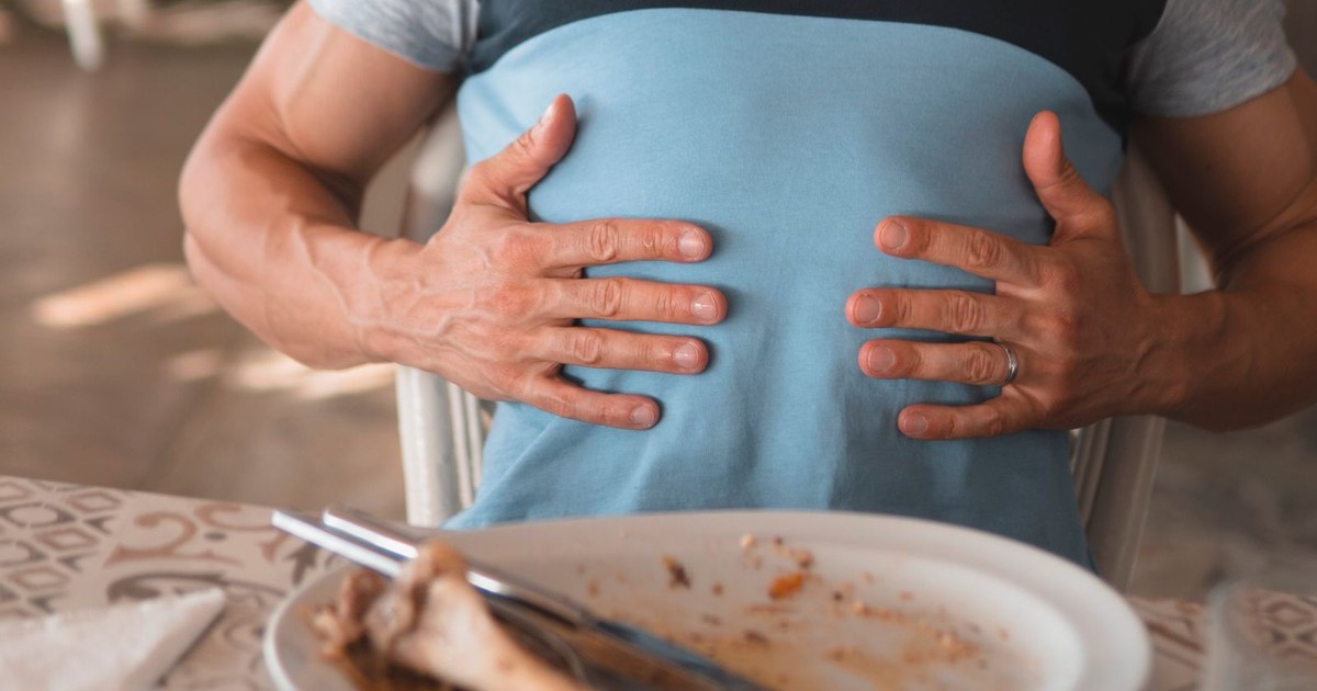 A seated man in a gray and blue color-block t-shirt rests both hands flat on his midsection at a dining table, with a finished plate in front of him