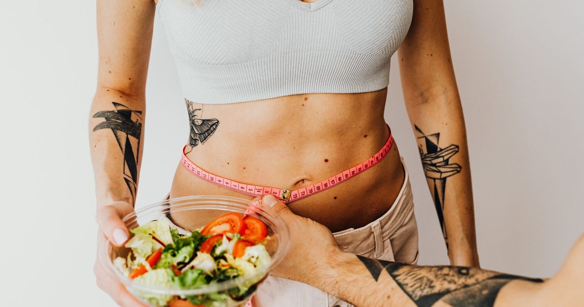 A woman measuring her waist while holding a bowl of fresh salad
