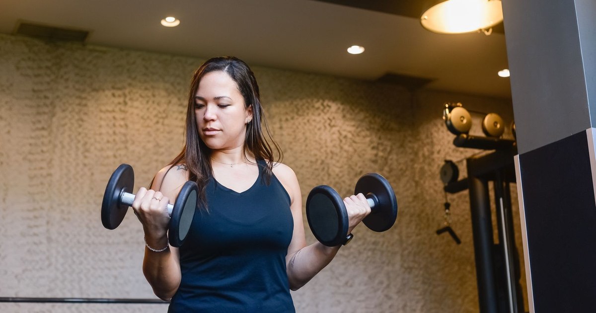 A woman in a black tank top curling two black dumbbells in a gym with a textured stone wall behind her