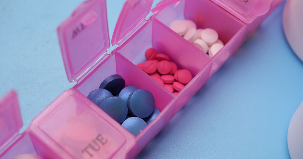 An open pink weekly pill organizer with colored tablets in each compartment on a blue surface