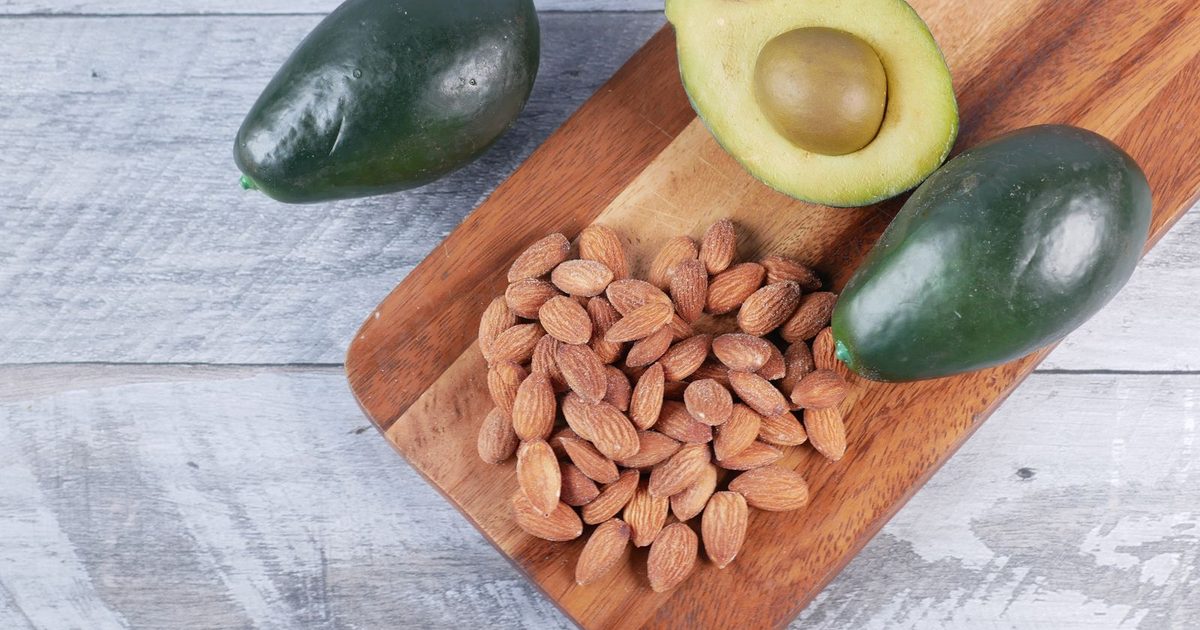 An overhead flat lay of a pile of whole almonds on a brown wooden serving board, with one halved avocado and two whole dark green avocados, set on a weathered gray and white wood surface