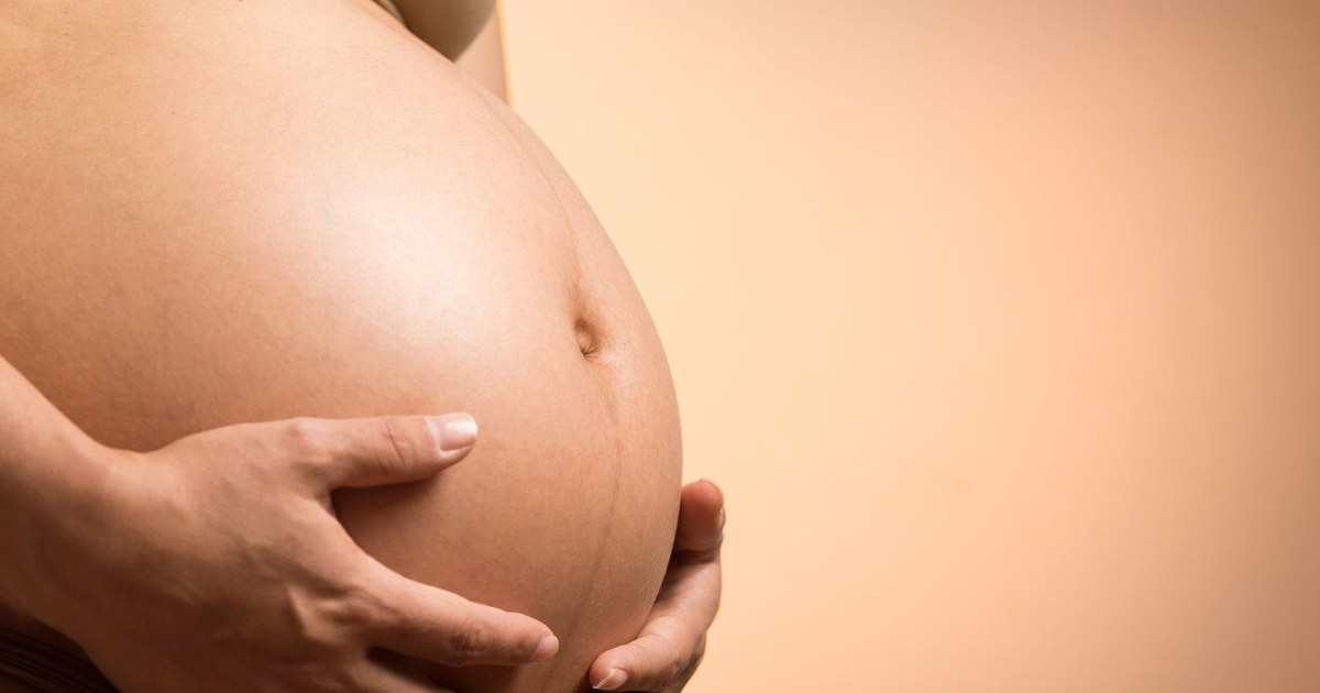A close-up of an expectant mother's bare pregnant belly cradled by both hands against a warm peach background, with no face visible
