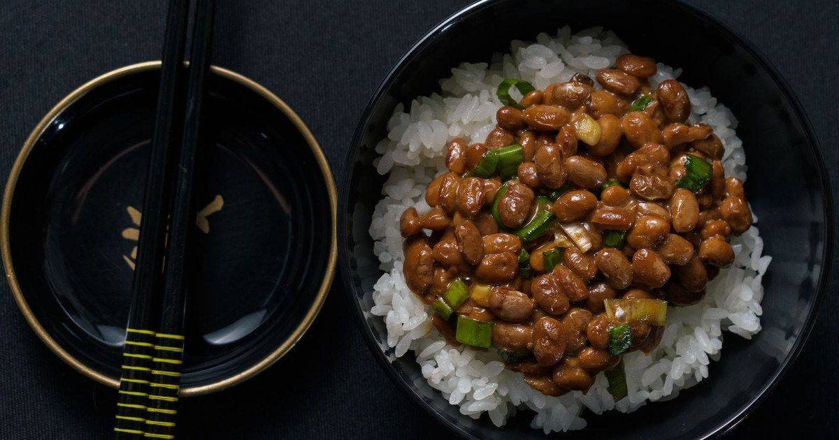 A bowl of natto fermented soybeans with scallions over white rice, beside chopsticks on a dark table
