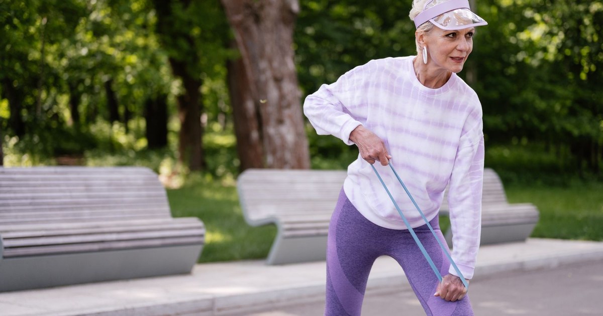 An older woman with short blonde hair in a lilac visor, lavender tie-dye sweatshirt and purple leggings leans forward while stretching a light blue resistance band with both hands on a paved path, with wooden park benches and sunlit green trees behind her