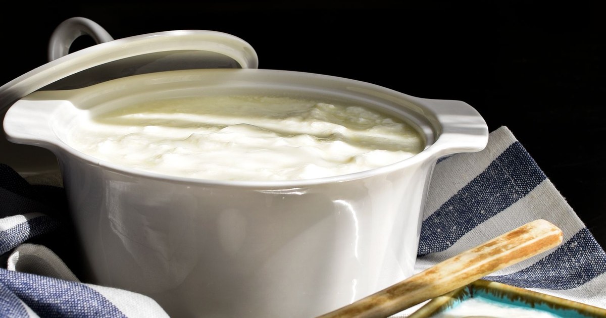 A white ceramic bowl of thick creamy plain yogurt on a blue and white striped cloth, with a wooden spoon handle at the lower right
