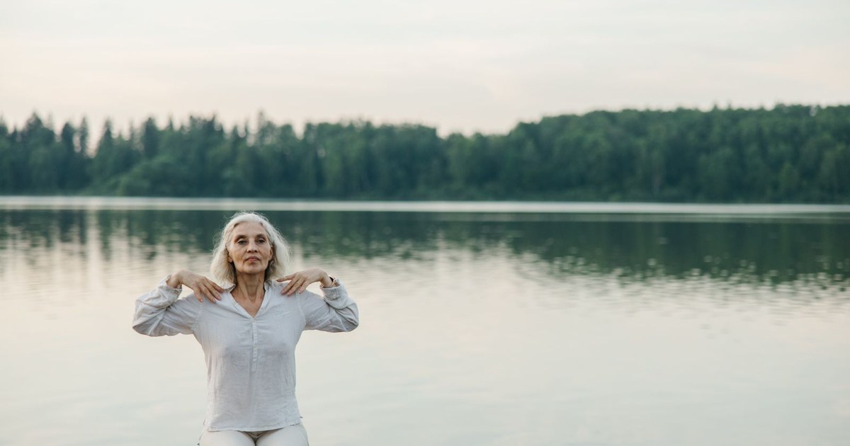 A woman with long white hair in a loose white linen shirt sits calmly at the edge of a still forest lake at dusk, resting her hands on her shoulders in a gentle stretch with a relaxed expression, dark green trees reflected in the water behind her