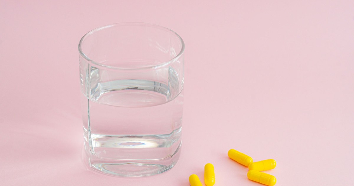 Yellow supplement capsules beside a glass of water on a pink background