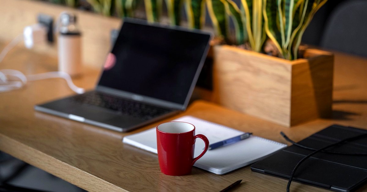 A tidy wooden desk in warm light with a coffee mug, an open notebook, a laptop, and a small plant