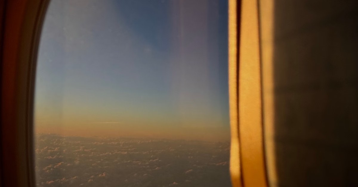 A view through an airplane cabin window at dusk, with a warm golden sky above a layer of clouds