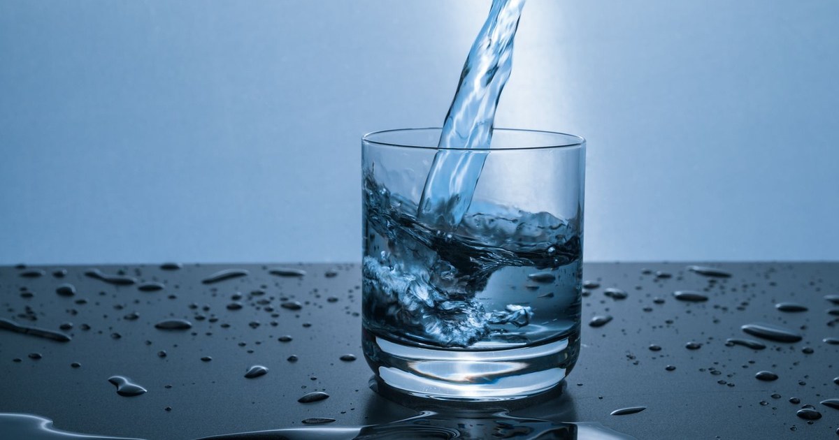 Water pouring into a clear drinking glass sitting on a wet dark surface scattered with droplets