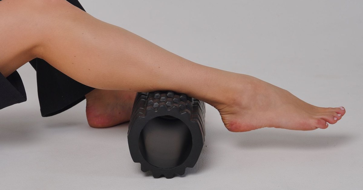 A person's bare lower leg resting on a black textured foam roller on a plain gray floor